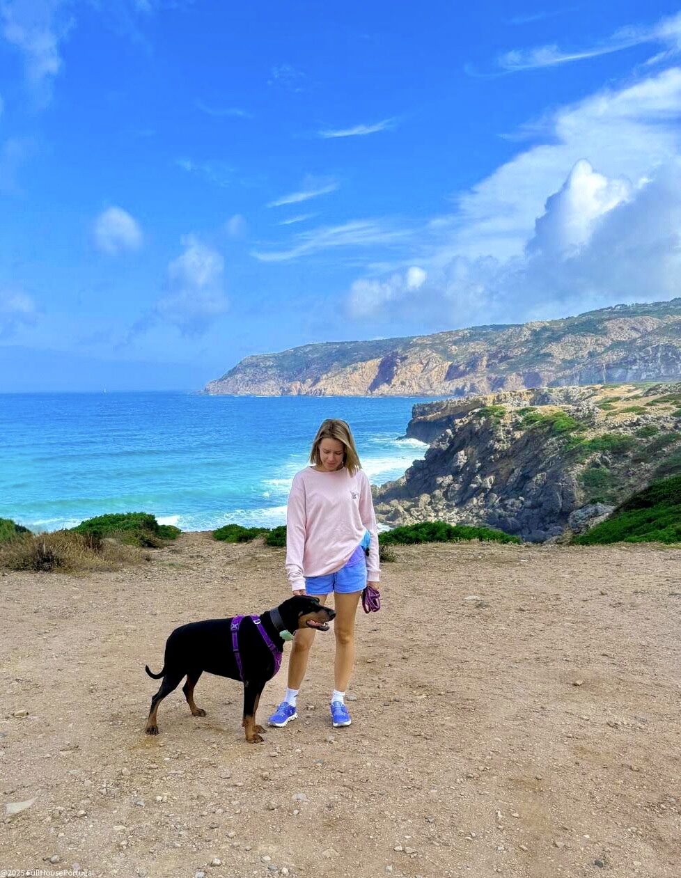 Kasia walking her dog on a clifftop trail above the Atlantic near Cascais, blue sea and rocky headland in the background