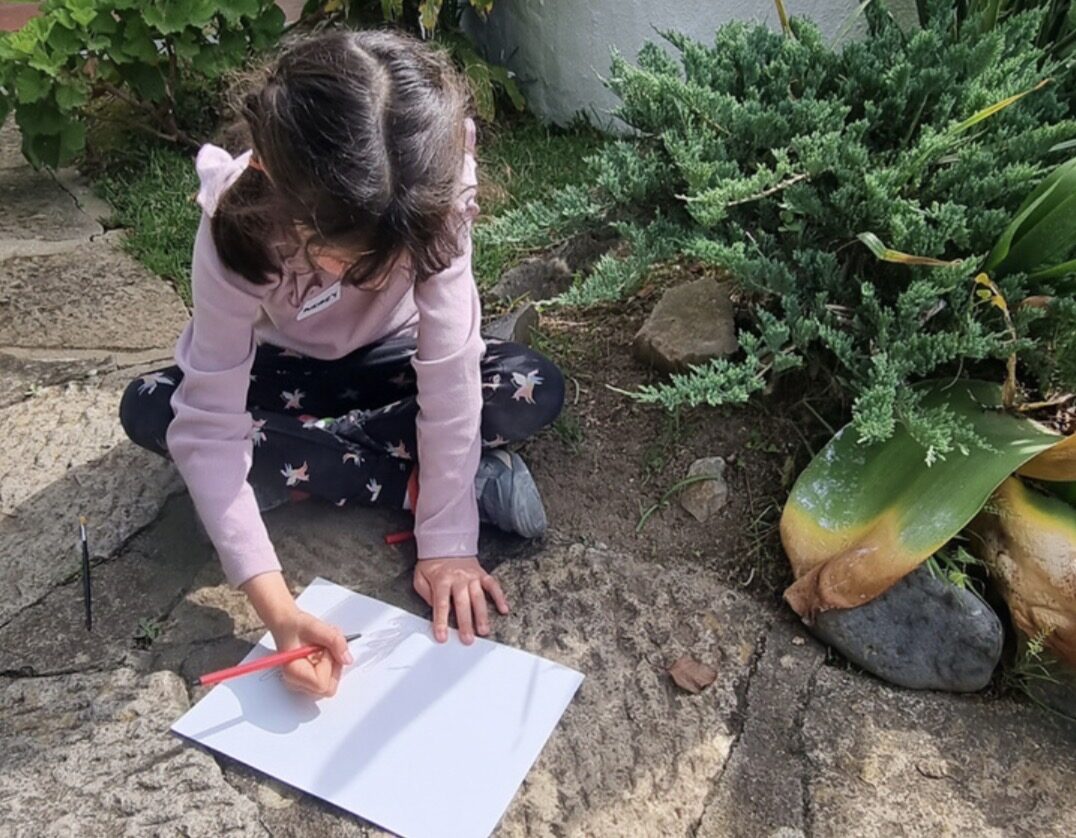 A child drawing on paper outside, sitting on stone steps surrounded by plants