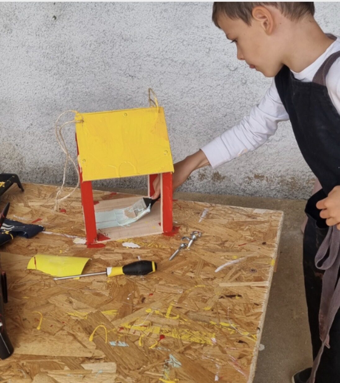 A child painting a small wooden birdhouse with a yellow roof and red posts during a woodworking session