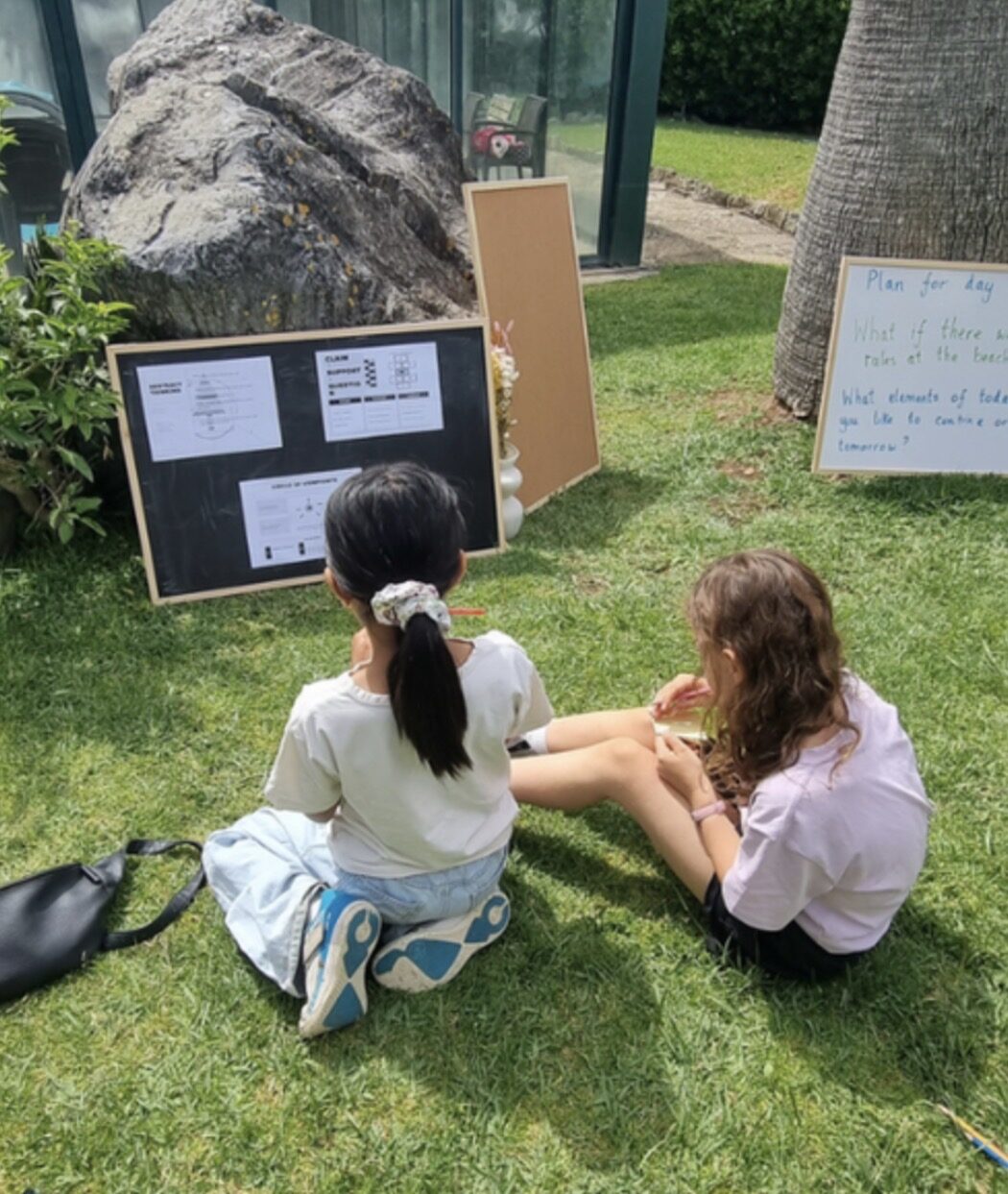 Two children sitting on the grass together, working on a shared project beside a pinboard with planning notes